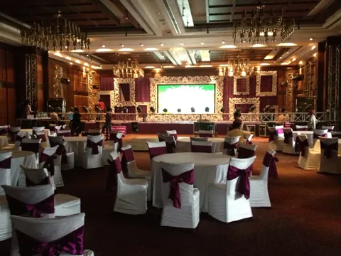 A banquet hall set with round tables and plum sashes facing an ornate white stage and LED wall