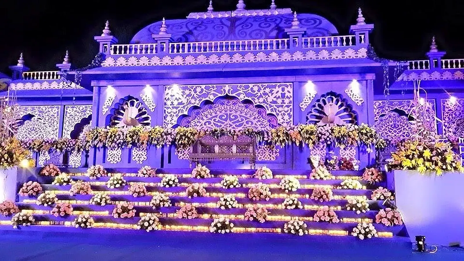 An illuminated wedding stage at night: a carved facade lit in lavender, peacock motifs and tiered steps banked with flowers, built by Mega Events in Mumbai